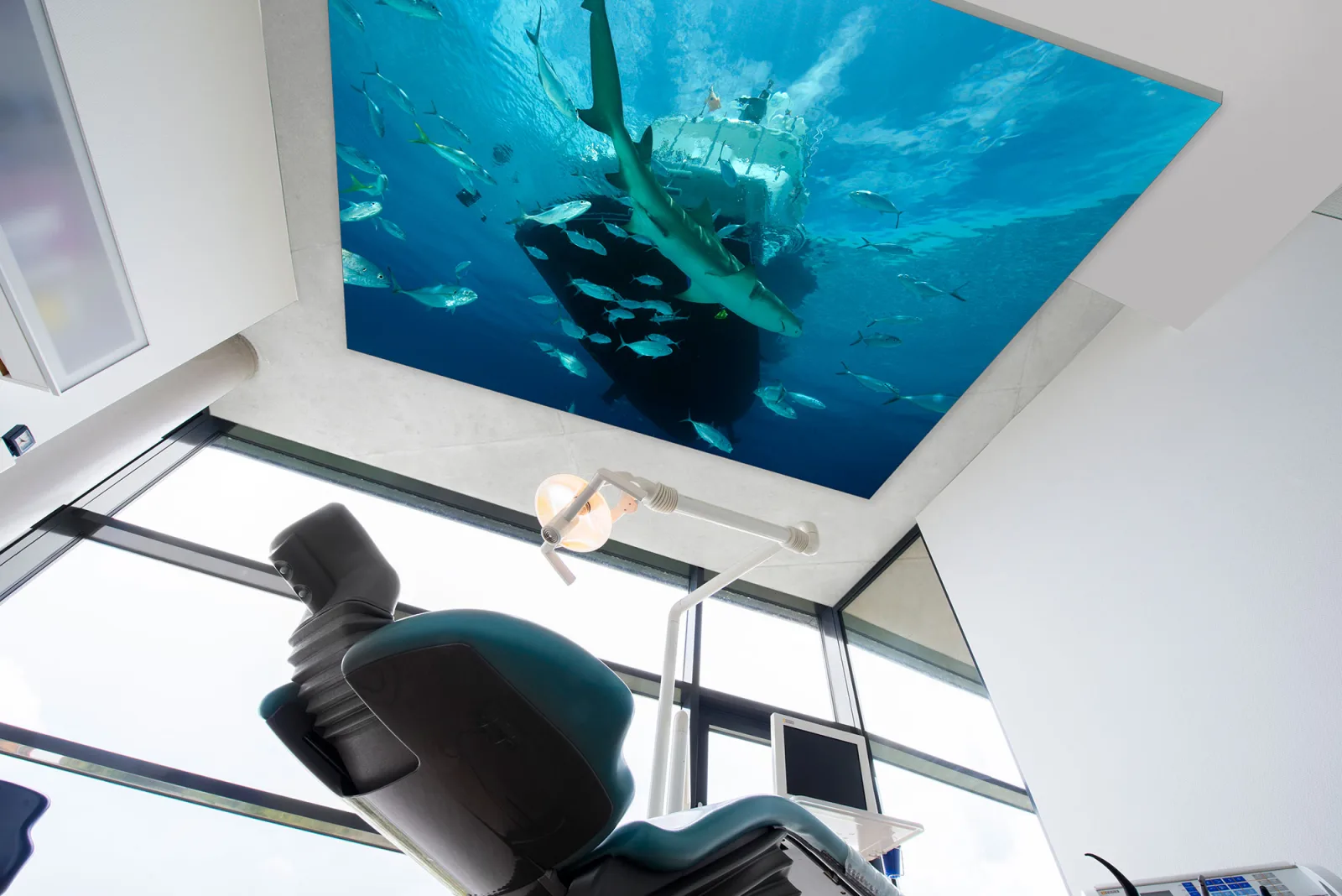 Blick vom Behandlungsstuhl nach oben auf eine hinterleuchtete Deckenfläche mit Unterwassermotiv, Fischschwarm und Hai über der Behandlungsleuchte