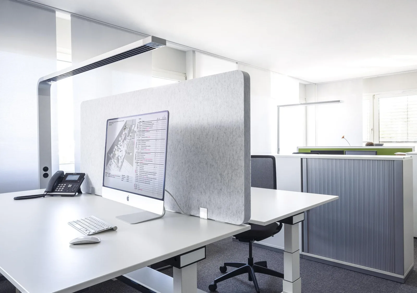Bildschirmarbeitsplatz mit schmaler Arbeitsplatzleuchte über dem Monitor, daneben eine graue Akustikstellwand und ein Rollladenschrank