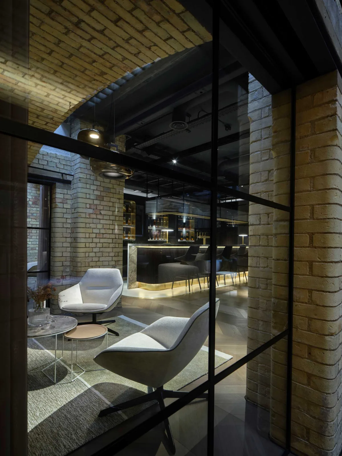 Hotellounge in einem Backsteingewölbe mit zwei hellen Loungesesseln, Beistelltischen und einer beleuchteten Bar dahinter