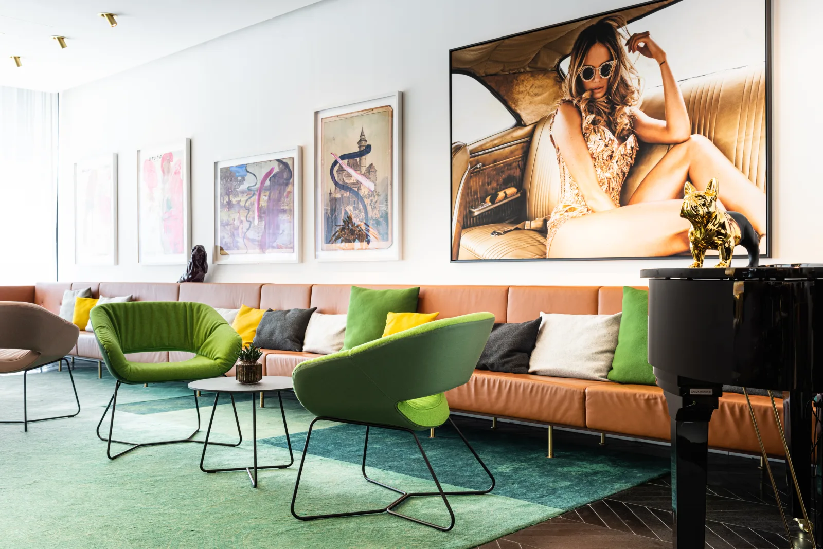 Hotellobby mit langer gepolsterter Sitzbank, grünen Sesseln und niedrigen Beistelltischen vor einer Bilderwand