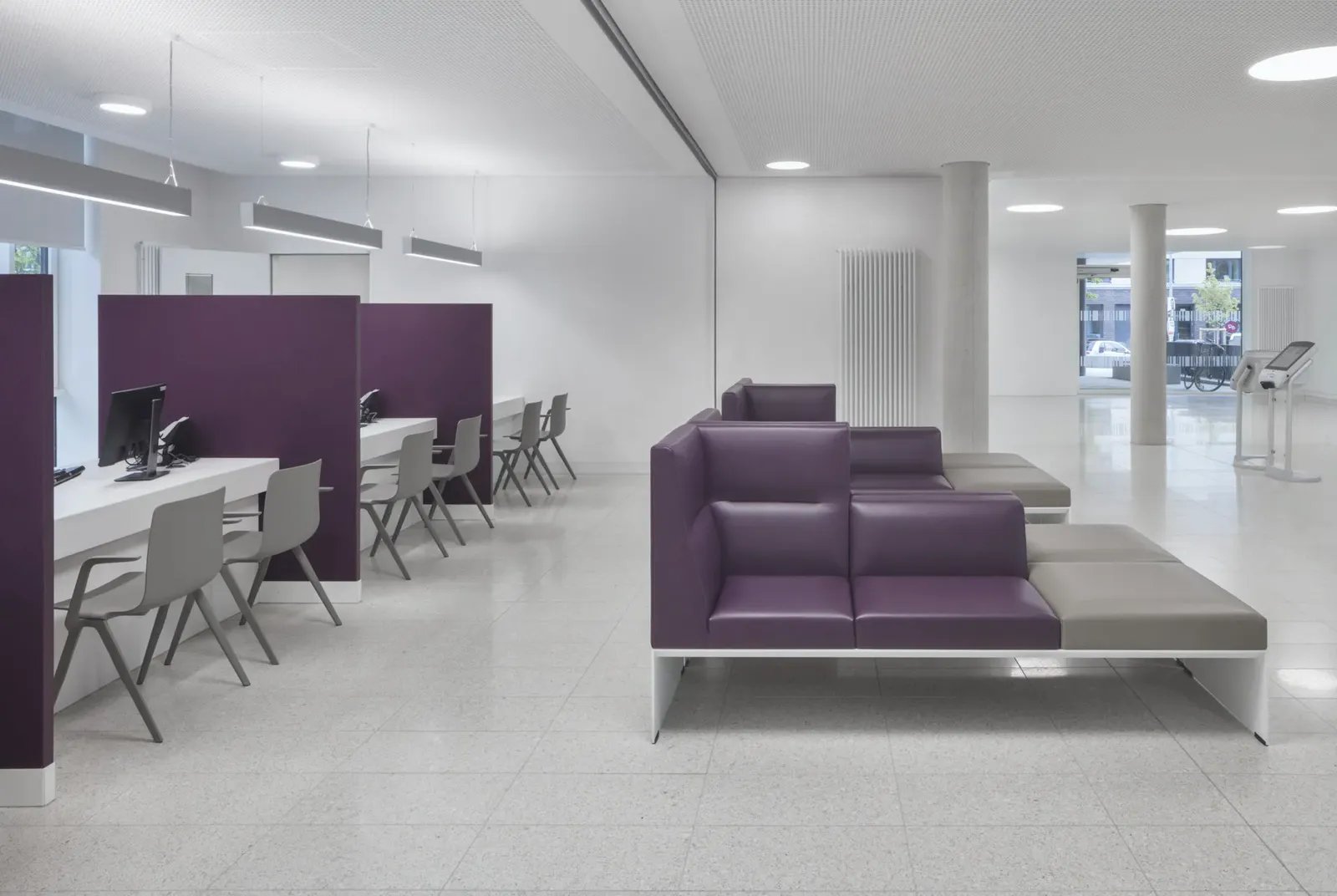 Patientenaufnahme mit mehreren Arbeitsplätzen nebeneinander, jeder durch eine violette Stellwand abgeschirmt, daneben eine Sitzinsel und ein Terminal