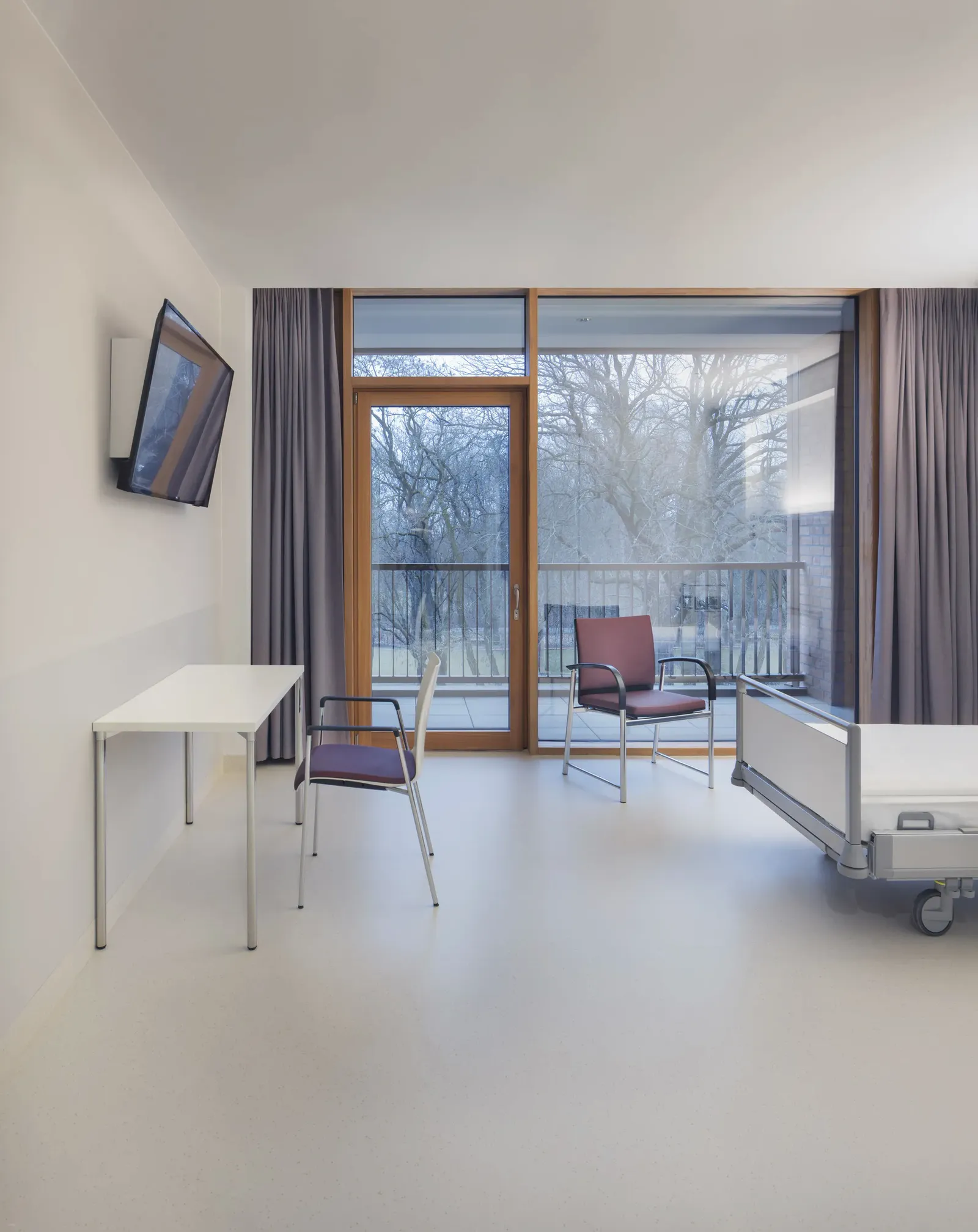Patientenzimmer mit Pflegebett, Tisch und zwei Stühlen vor der Fensterfront, Fernseher an der Wand