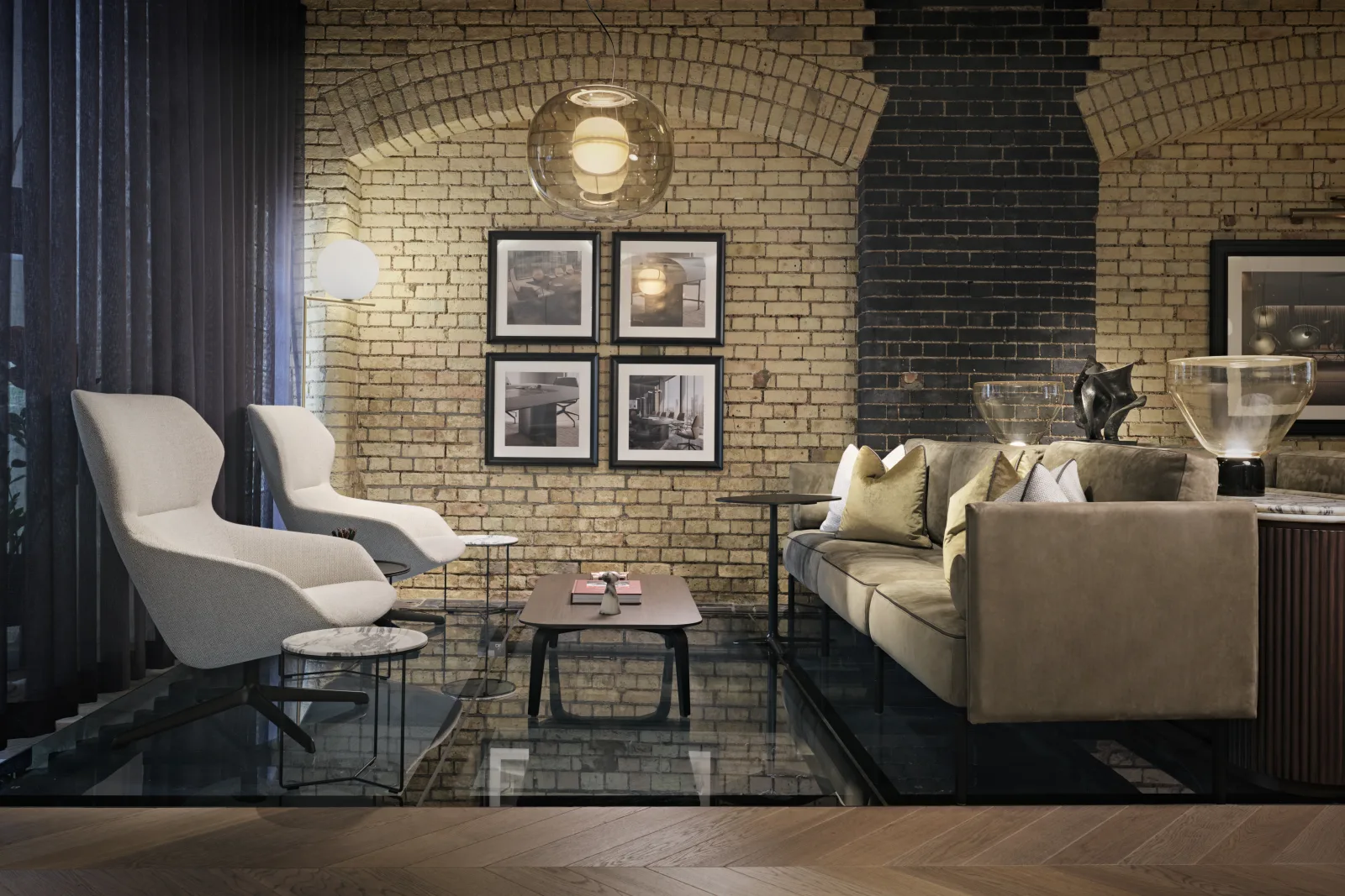 Lounge mit zwei hellen Sesseln, Sofa und Beistelltischen vor einer Backsteinwand mit Bildern und Pendelleuchte