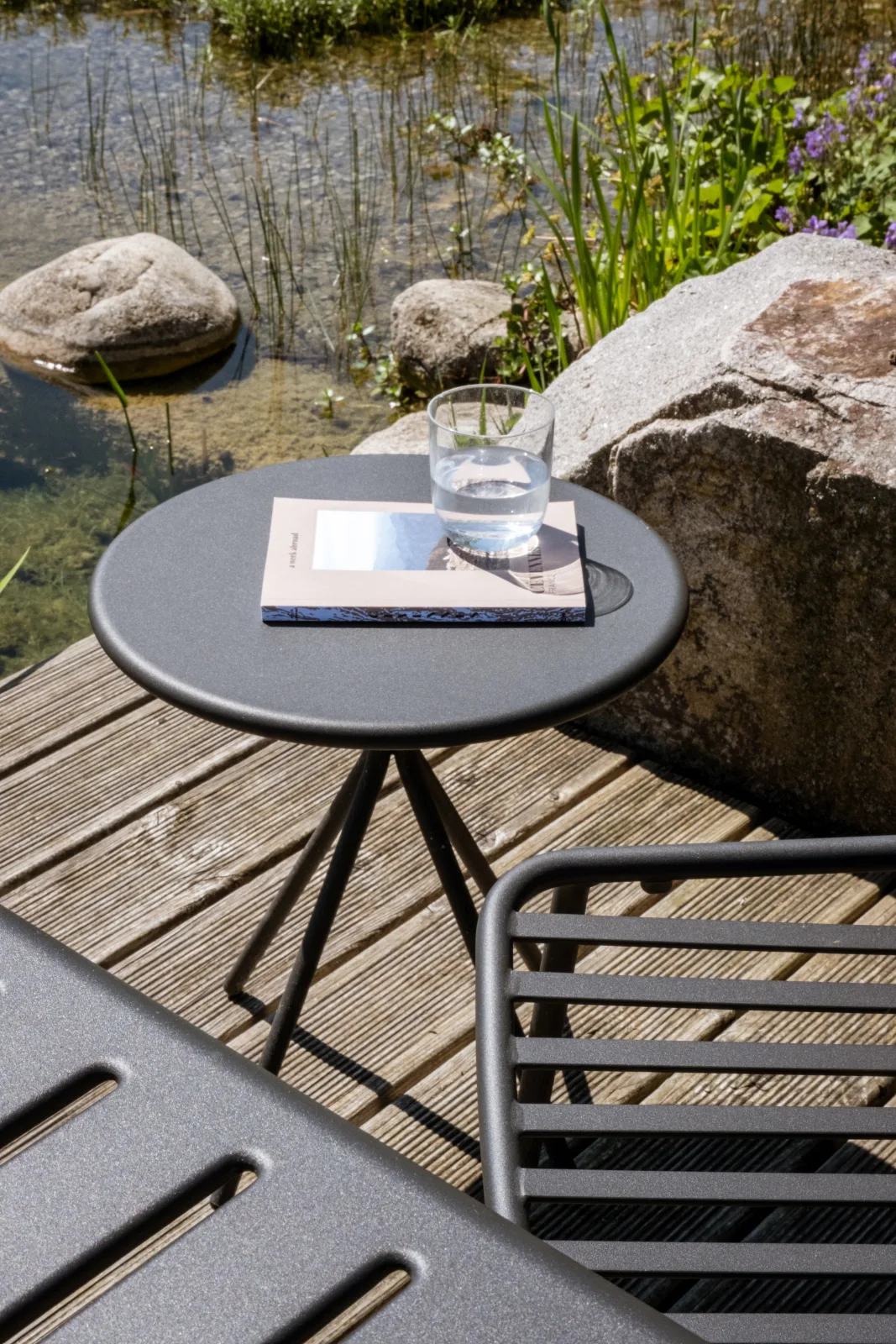 Runder Beistelltisch mit Glas und Buch auf einem Holzdeck, daneben eine Stuhllehne und eine Tischkante