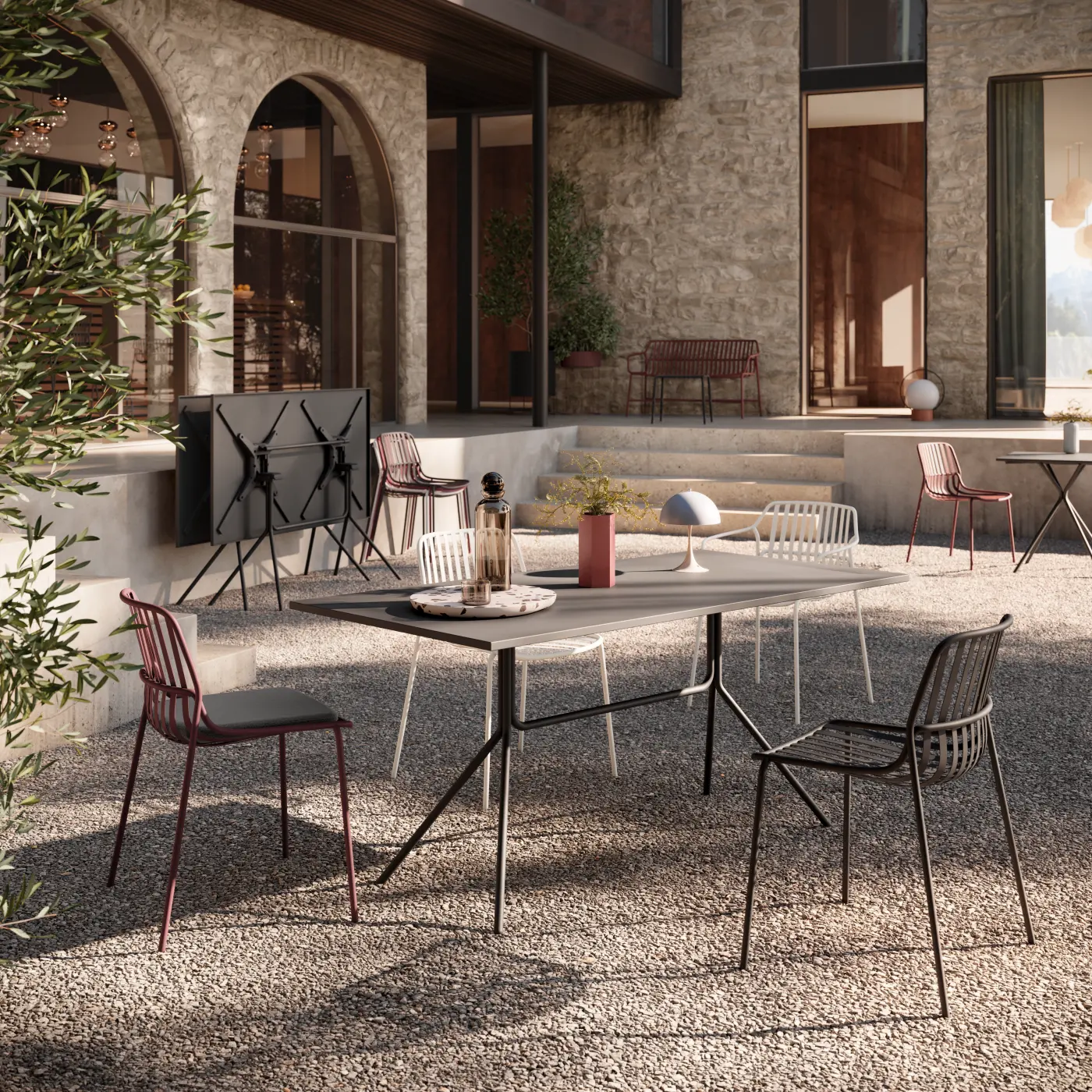 Terrasse eines Steinhauses mit Rundbögen, grauem Tisch, weißen und bordeauxroten Drahtstühlen und Olivenbaum