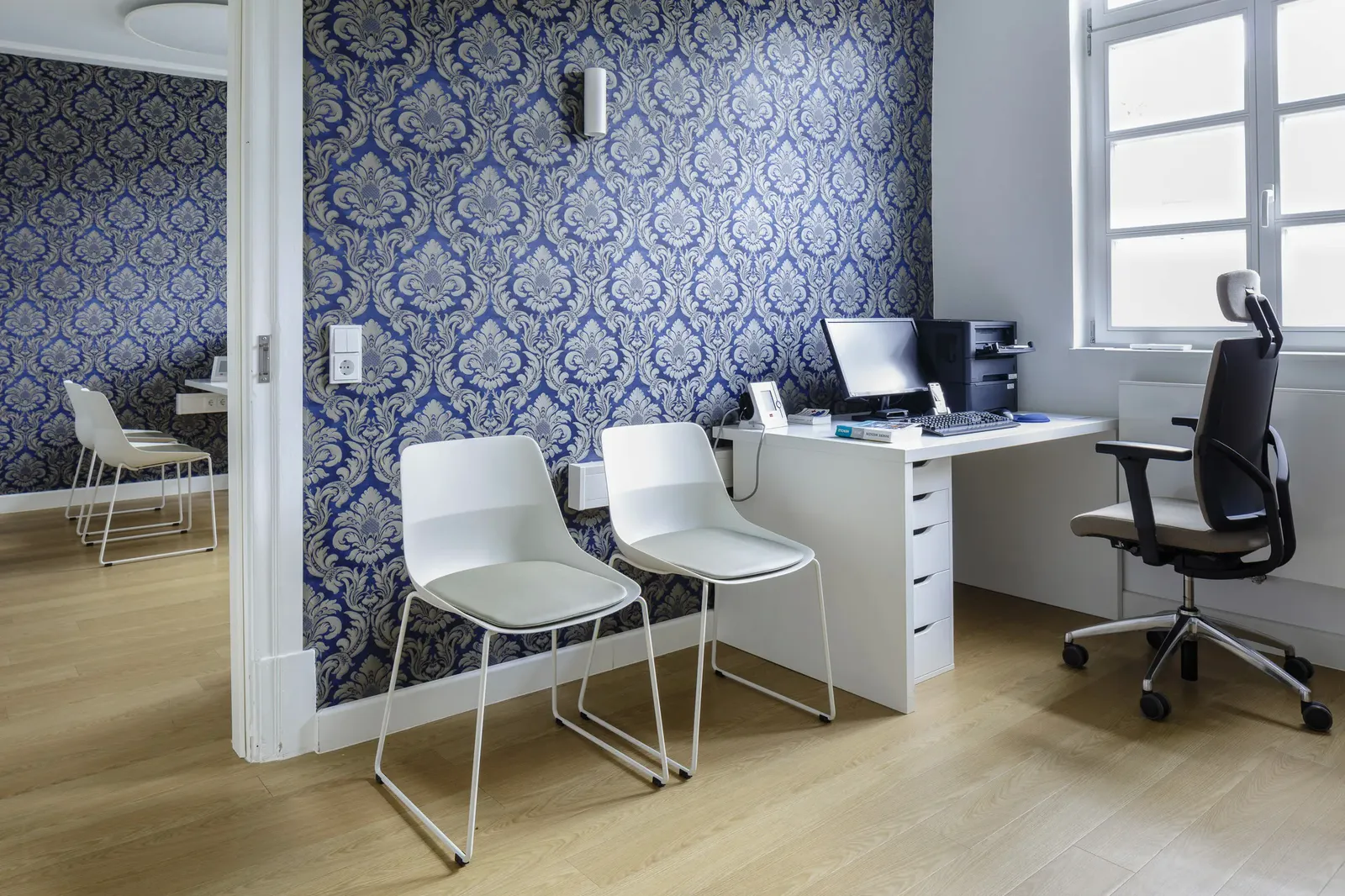 Sprechzimmer einer Arztpraxis mit Schreibtisch, Drehstuhl und zwei weißen Besucherstühlen vor gemusterter Wand