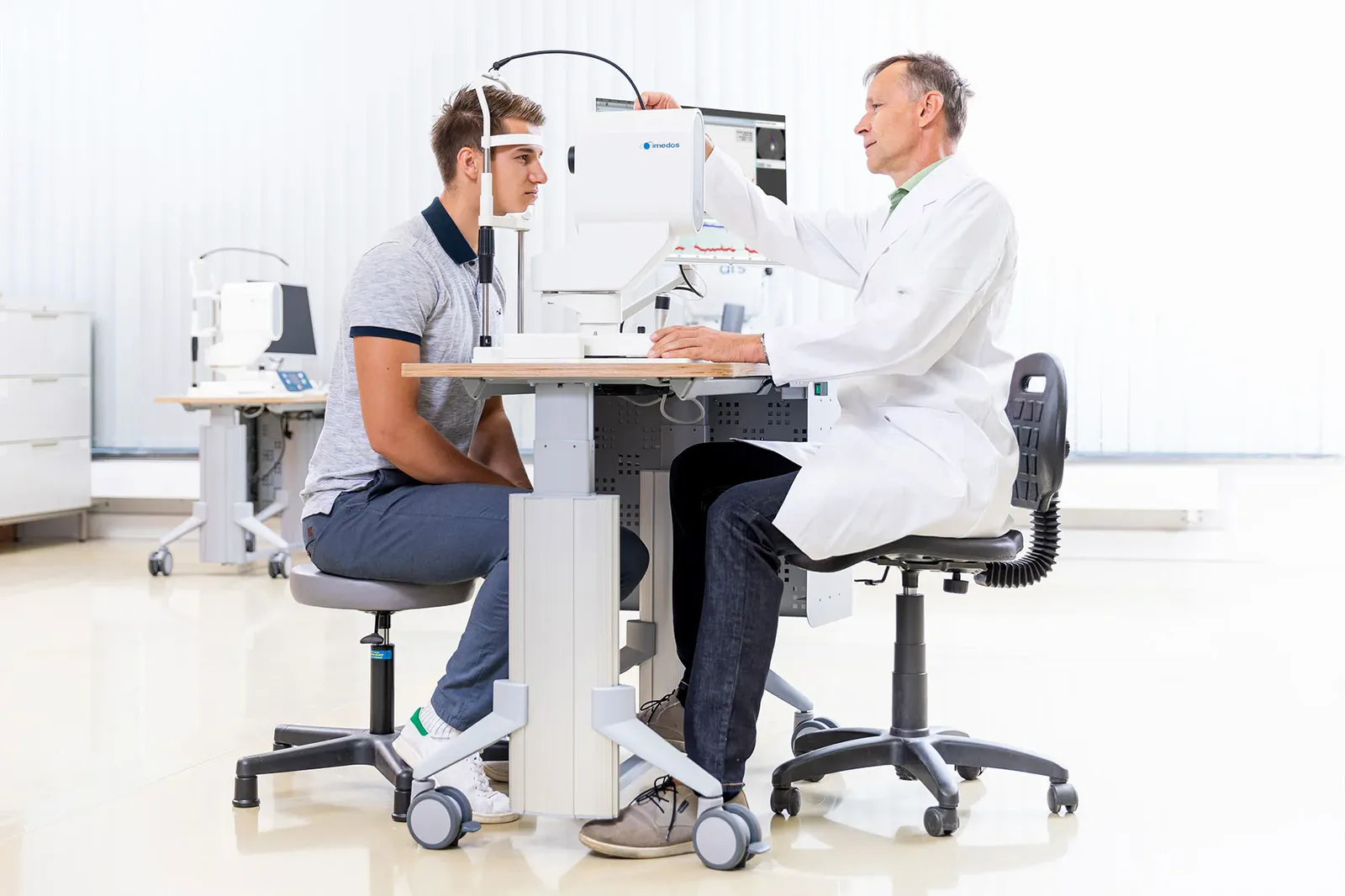 Ärztin und Patient sitzen einander an einem höhenverstellbaren Diagnostiktisch gegenüber, beide auf Rollhockern
