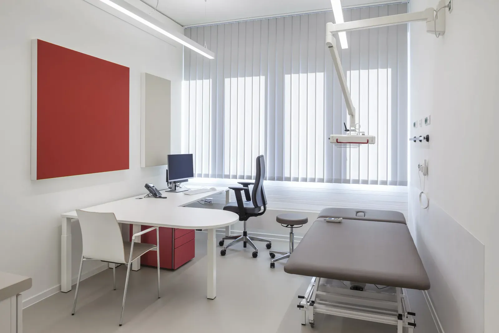 Sprechzimmer mit Schreibtisch, Bildschirm und Drehstuhl, am Rand eine Untersuchungsliege vor einer roten Akustikfläche