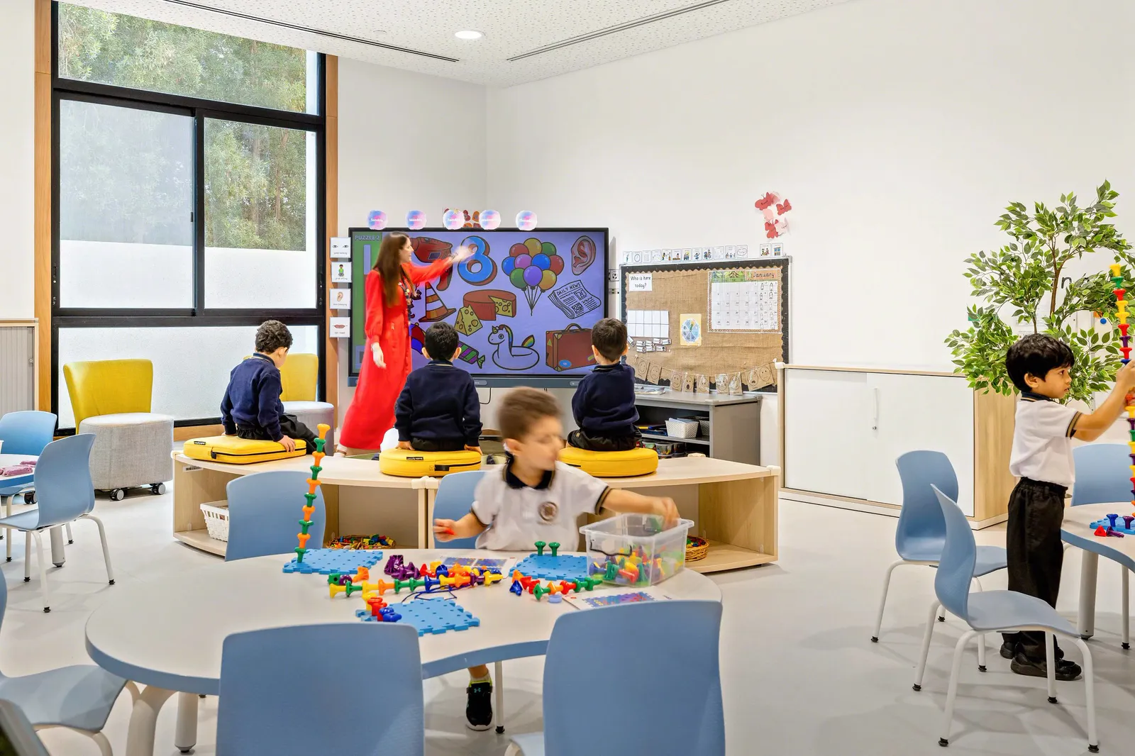 Unterrichtsraum mit runden Gruppentischen, blauen Schulstühlen und einer Lehrkraft am digitalen Whiteboard