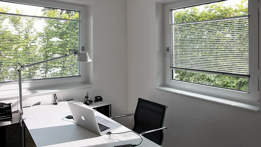 Helles Arbeitszimmer mit weißem Schreibtisch, Laptop und schwarzem Drehstuhl, an beiden Fenstern silberne Jalousien